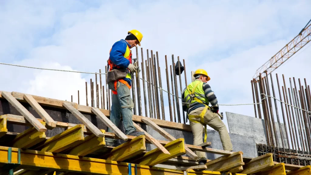 Trabalhadores da construção civil em atividade sobre estrutura de concreto com vergalhões aparentes, avaliando a escolha entre estrutura metálica ou concreto.