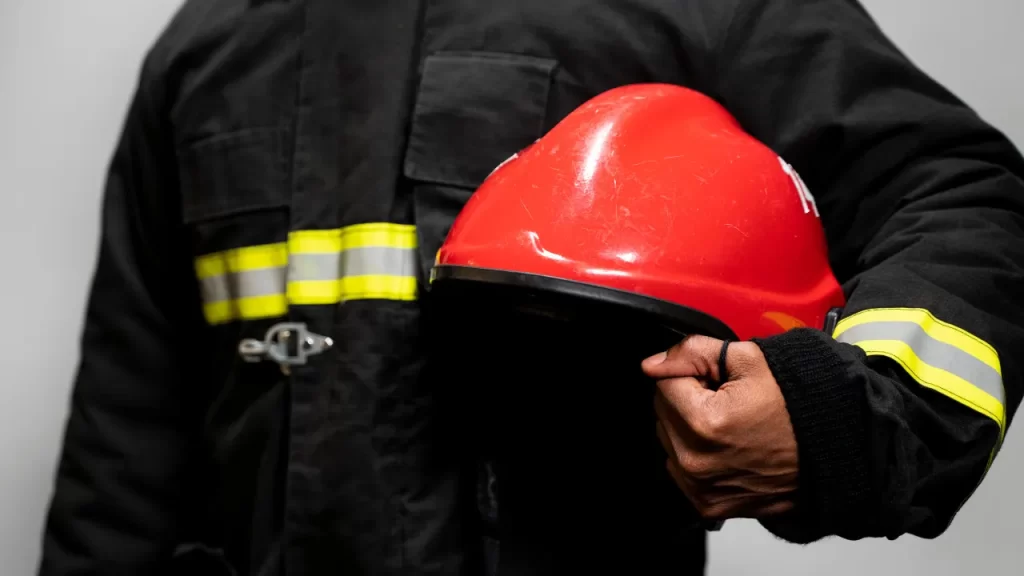 Bombeiro em uniforme com faixas refletivas segurando capacete vermelho, representando conformidade com os Critérios do Corpo de Bombeiros para Aprovação.